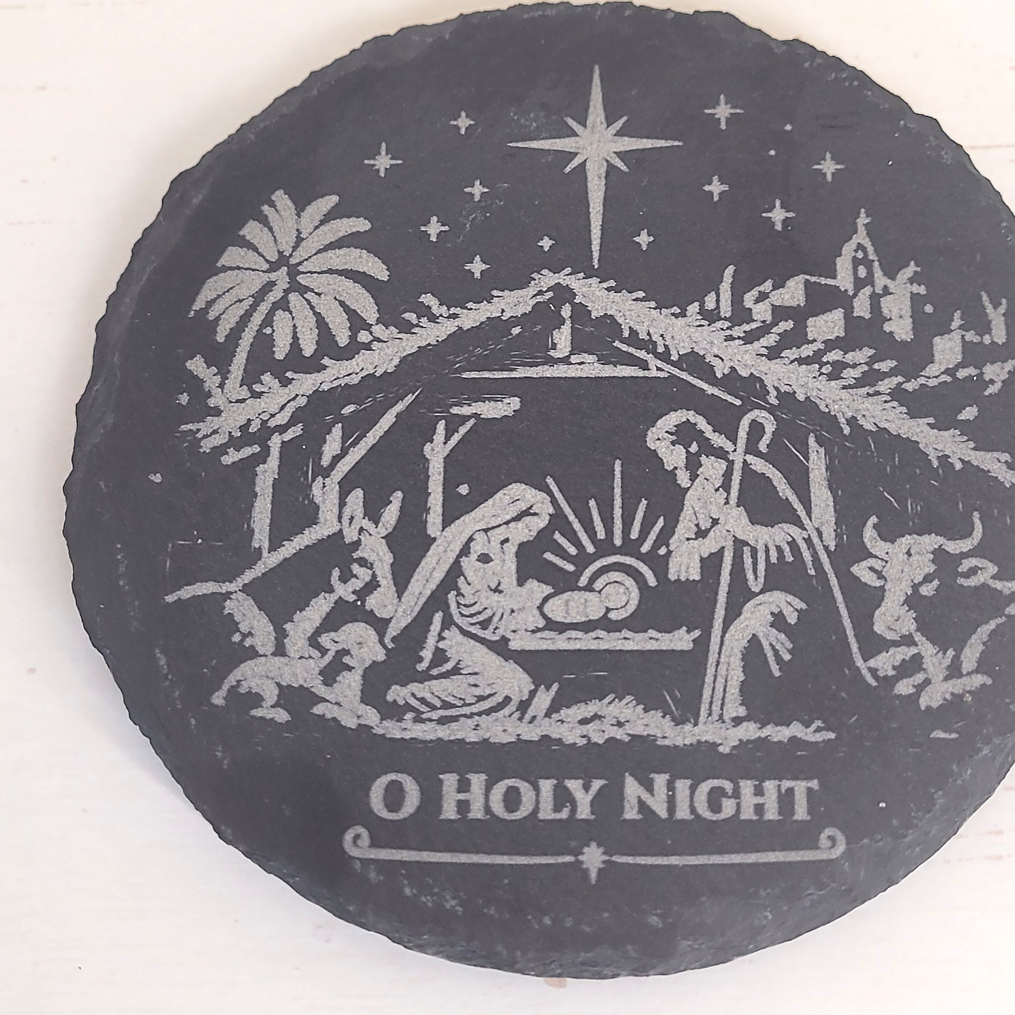 One round slate coaster seen from above, engraved with a nativity scene under a bright star and the words "O Holy Night". The set contains four identical coasters.